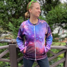 Load image into Gallery viewer, Emily wearing the NGC 602 light jacket, seen from the front looking to the side with a weird smile with her hands in the front pockets, with a fence and greenery in the background