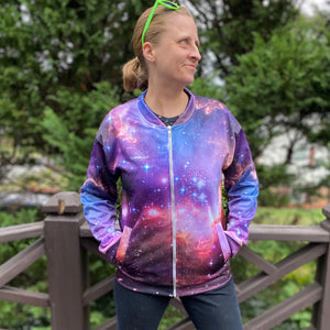 Emily wearing the NGC 602 light jacket, seen from the front looking to the side with a weird smile with her hands in the front pockets, with a fence and greenery in the background