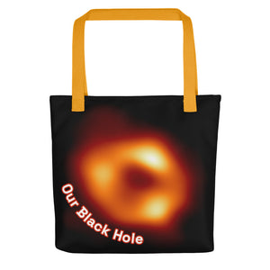 Digital mock up of a black tote bag with yellow handle printed with the Event Horizon Telescope image of the Sgr A* supermassive black hole in the Milky Way Galaxy with curved text reading "Our Black Hole" in red and white on one side.