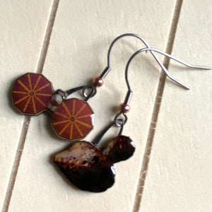 Mismatched earrngs made from upcycled paperboard, one an illustration of the Lucy mission soacecraft and the other binary asteroids in partial shadow, shown reating on a light striped surface.