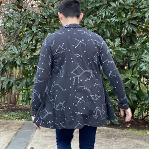 Seen from behind, Matthew wears a dark gray cardigan with white constellation stars and lines, mid-stride with arms away from his sides and the cardigan flaring out a bit, with green foliage in the background.