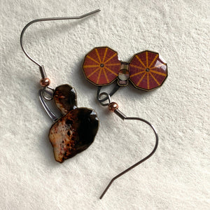 Mismatched earrngs made from upcycled paperboard, one an illustration of the Lucy mission soacecraft and the other binary asteroids in partial shadow, shown on a textured white earring display on a textured white background.