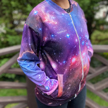 Load image into Gallery viewer, Emily wearing the NGC 602 light jacket, shown from the front/side with her hands in the front pockets, with an out-of-focus fence and greenery in the background