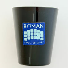 Load image into Gallery viewer, A black shot glass featuring a blue and white sticker that reads 'ROMAN SPACE TELESCOPE' with a grid design, set against a plain white background.