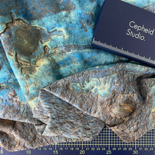Load image into Gallery viewer, A close-up of a Mars surface image fabric sample with a mottled blue, teal, and brown organic pattern, laid out on a gridded cutting mat with a dark blue 'Cepheid Studio.' box placed on top.