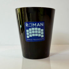 Load image into Gallery viewer, A black glossy shot glass featuring a blue and white sticker that reads 'ROMAN SPACE TELESCOPE' with a grid logo, set against a plain white background.
