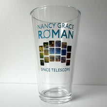 Load image into Gallery viewer, A clear glass pint glass featuring the text 'NANCY GRACE RÓMAN SPACE TELESCOPE' in blue, with a central graphic of colorful square windows resembling the James Webb Space Telescope's views, set against a plain light gray background.