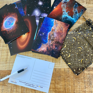 JWST First Year postcards arranged on a brown wood surface, including one flipped to the back, a STARtorialist pen, and a black and gold organza bag
