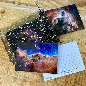 JWST First Year postcards arranged on a brown wood surface, including one flipped to the back, and a black and gold organza bag