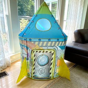 Play rocketship tent set up in the corner of a living room by a sunny window, shown with the flap closed.