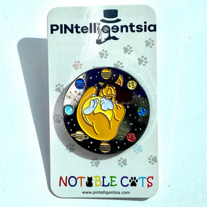 Cat-purr-nicur round enamel pin with an orange cartoon cat holding the Sun surrounded by planets on a black space background on a paper card