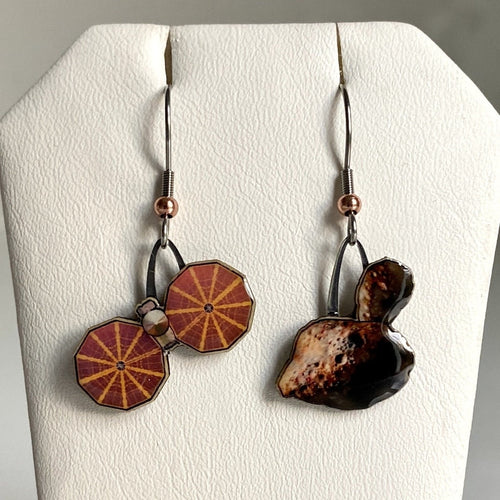 Mismatched earrngs made from upcycled paperboard, one an illustration of the Lucy mission soacecraft and the other binary asteroids in partial shadow, shown hanging on a textured white earring display on a white background.