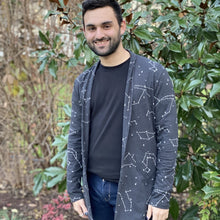 Load image into Gallery viewer, Matthew wears a dark gray cardigan with white constellation stars and lines over a dark t-shirt, smiling with hands at his sides, with green foliage and brown branches in the background.